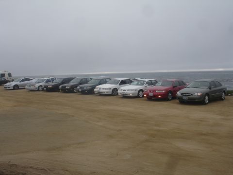1990 Ford Taurus - A line of sedans are parked on a dirt lot with the ocean in the background.