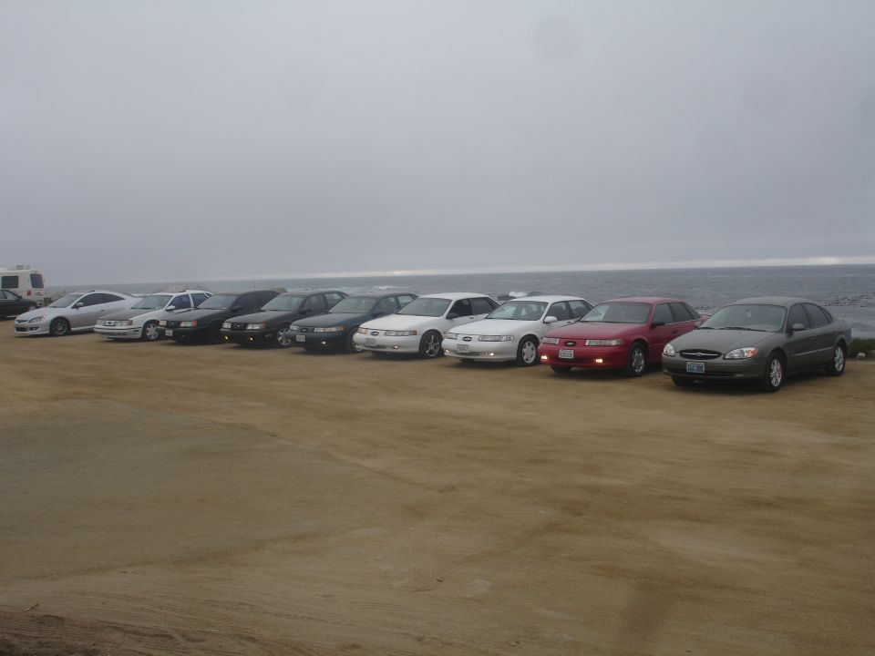 several sedans parked in a row on a dirt lot facing the ocean