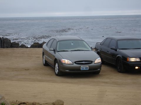 1990 Ford Taurus - A gray sedan is parked on a sandy beach with the ocean in the background, alongside another dark sedan.