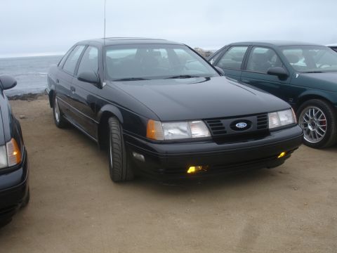 1990 Ford Taurus - A black 1990 Ford Taurus sedan is seen parked near the coast, with another vehicle visible to its right.