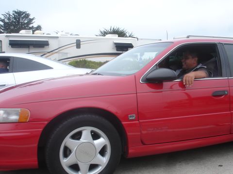 1990 Ford Taurus - Man driving a red 1990 Ford Taurus sedan, with a white car parked nearby.