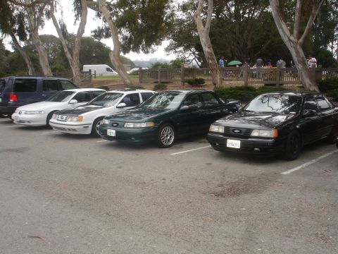 1990 Ford Taurus - Several Ford Taurus sedans are parked in a lot, with trees and people in the background.