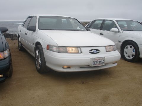 1990 Ford Taurus - Two white Ford Tauruses are parked near the coast under an overcast sky.