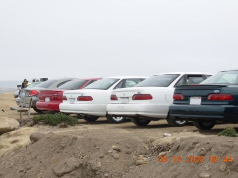1995 Ford Taurus - A line of sedans, including a 1995 Ford Taurus, are parked on a dirt lot near the coast.