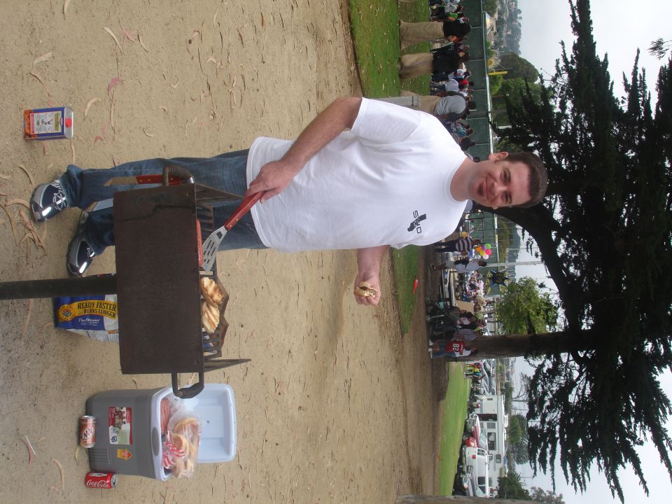 Man in white t-shirt grilling hot dogs at a park event, with a cooler and drinks nearby