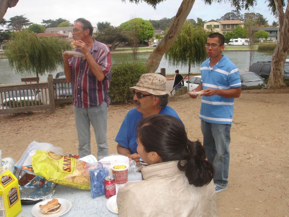Four people at a picnic table with food and drinks, a lake and trees in the background