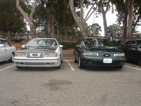 1990 Ford Taurus - Two Ford Taurus sedans are parked in a lot, with trees and a wooden fence in the background.