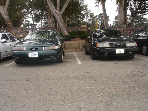 1990 Ford Taurus - Two Ford Taurus sedans are parked in a lot with trees and a wooden fence in the background.