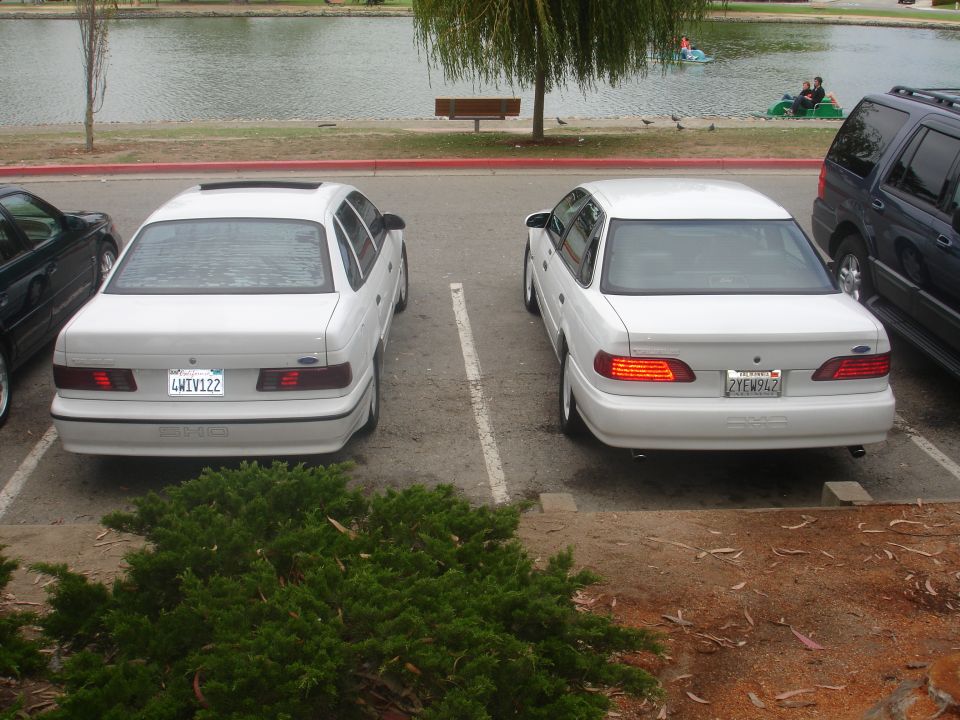 Two white sedans parked next to each other, one with a sunroof. A dark SUV is parked to the right. Lake and people in boats in background