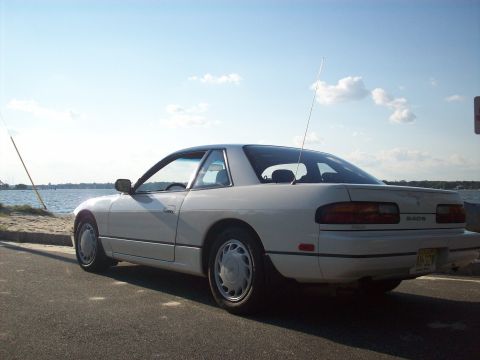 1990 Nissan Sentra - Rear view of a white 1990 Nissan 240SX coupe with a spoiler, parked near a body of water under a partly cloudy sky.