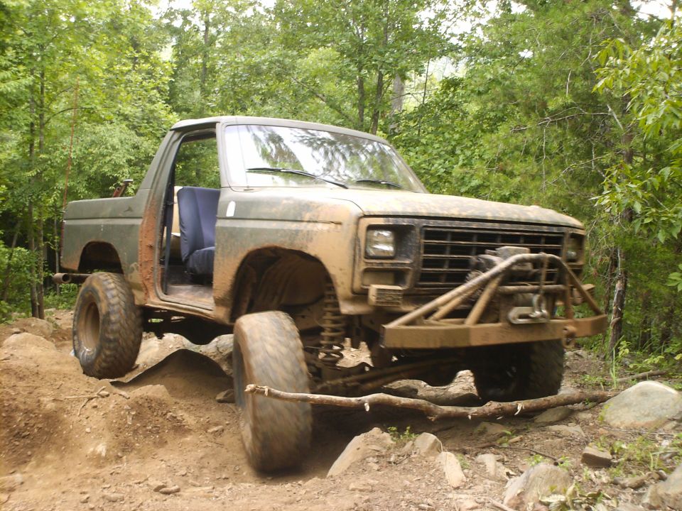 Muddy 1980 Ford Bronco off-roading on a rocky trail with the driver's door open.