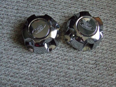 2003 Ford Crown Victoria - Two chrome Ford hubcaps are shown on a carpeted surface. The Ford logo is visible on each hubcap.