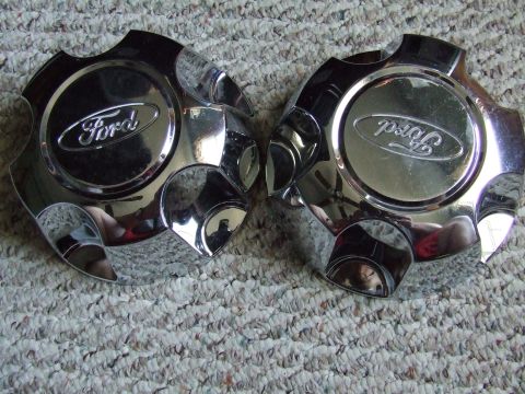 2003 Ford Crown Victoria - Two chrome Ford center caps with the Ford logo are shown resting on a carpeted surface. The center caps have a multi-pointed design.