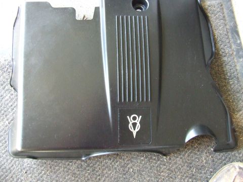 2003 Ford Crown Victoria - A black plastic engine cover with a V8 emblem is shown from a top-down angle. The cover has vertical ribbed detailing on the right side.