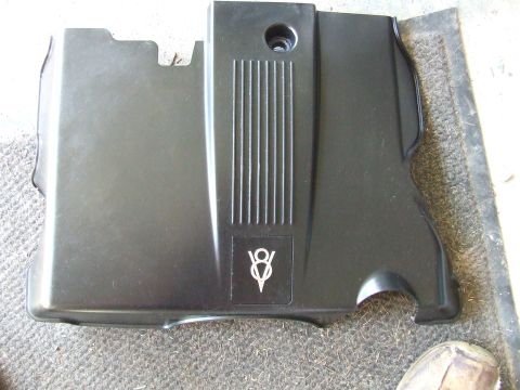 2003 Ford Crown Victoria - A black plastic engine cover with a stylized "V8" logo is shown from a top-down perspective, resting on a textured surface.