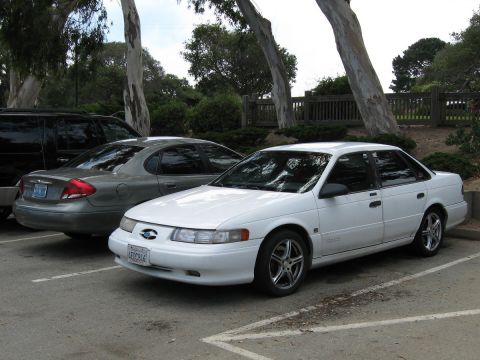 2003 Ford Taurus - Front three-quarter view of a white second-generation Ford Taurus SHO sedan on aftermarket five-spoke chrome wheels, parked in a lot under…