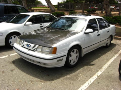 2003 Ford Taurus - A white four-door sedan with a carbon fiber hood is parked in a lot. The car has a dark tinted sunroof and is angled towards the front.