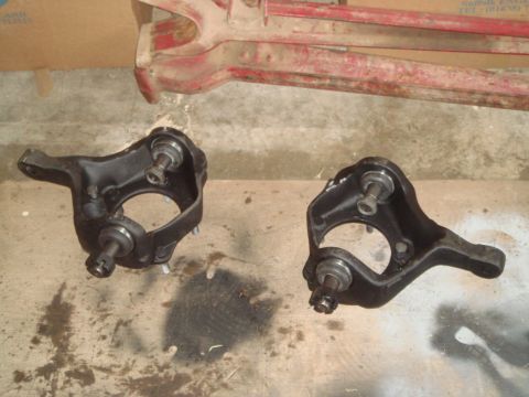 1992 Ford Bronco - Two black painted steering knuckles with ball joints and hardware are laid out on a concrete floor. A red metal suspension component is vis…