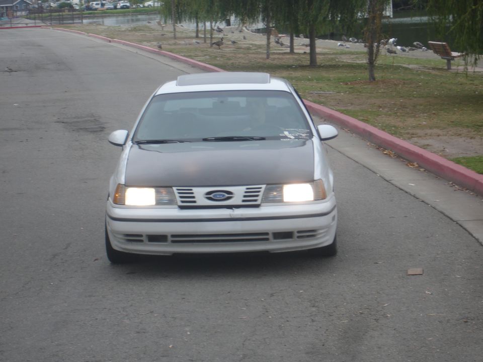 White Ford Taurus with a dark hood driving down a road, headlights on.