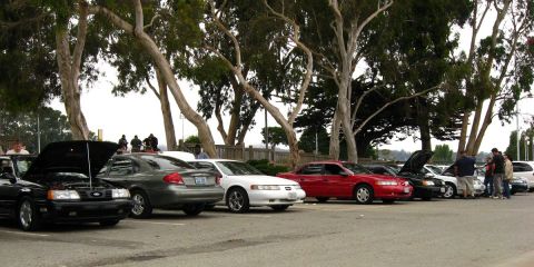 2003 Ford Taurus - A line of sedans are parked under trees, with several people standing around them. The hood of the black car on the left is open, revealing…