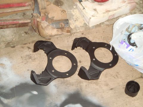 1992 Ford Bronco - Two black metal brake caliper brackets are laid out on a concrete floor. A plastic bag filled with parts and a small black plastic cap are…