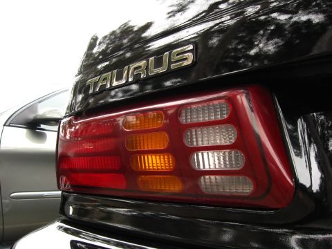 2003 Ford Taurus - Close-up of the rear corner of a glossy black Ford Taurus showing the chrome "TAURUS" trunk-lid badge above a multi-segment tail lamp with…
