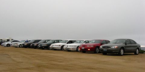 2003 Ford Taurus - Nine Ford Taurus and Mercury Sable sedans from multiple generations lined up nose-out on a sandy coastal lot under heavy overcast, with a g…