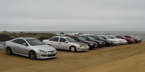 2003 Ford Taurus - A line of sedans and a coupe are parked on a sandy beach with the ocean and rocky shoreline in the background under an overcast sky.