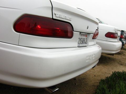 2003 Ford Taurus - Low-angle close-up of the rear corner of a white Ford Taurus sedan, showing the red wraparound taillight, chrome "TAURUS" badge and a Calif…