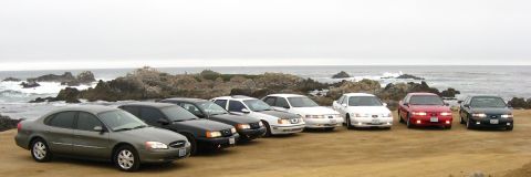 2003 Ford Taurus - Panoramic lineup of eight Ford Taurus sedans angled nose-out on a dirt bluff above a rocky surf-washed coastline, ranging from a gray 2000s…