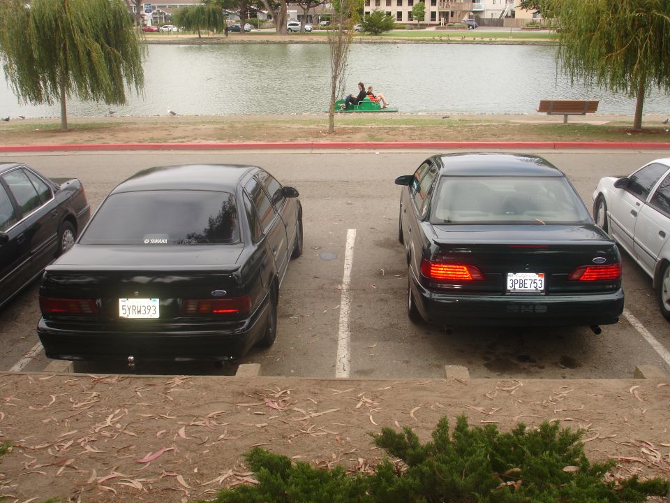 Two black sedans parked in a lot, one with license plate 3PBE753, with a lake in the background.