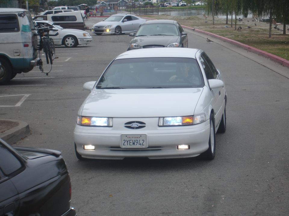 White 1990s Ford Taurus with headlights on, facing forward.