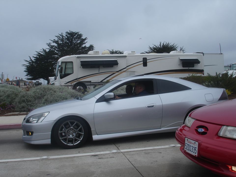 Silver coupe and red car parked on street with RV in background.