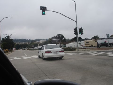 1990 Ford Taurus - A white sedan is seen from the rear, driving on a road with other vehicles under a cloudy sky.