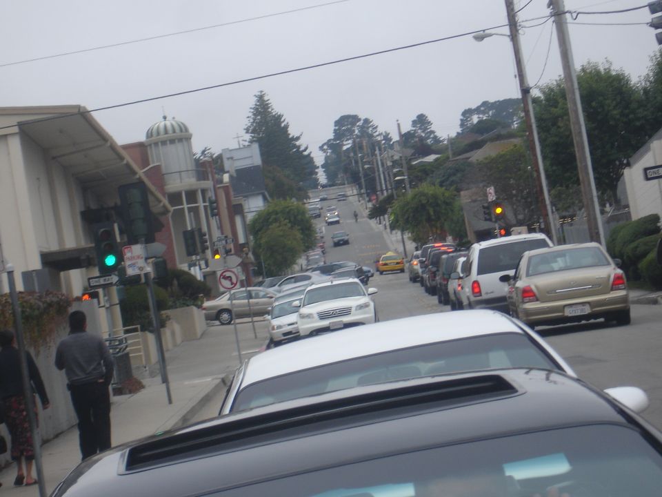 Gold sedan in traffic on a street with buildings and trees.