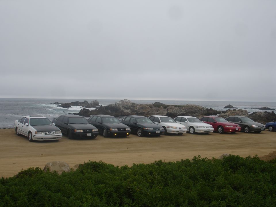 A line of sedans parked on a dirt lot by the ocean.