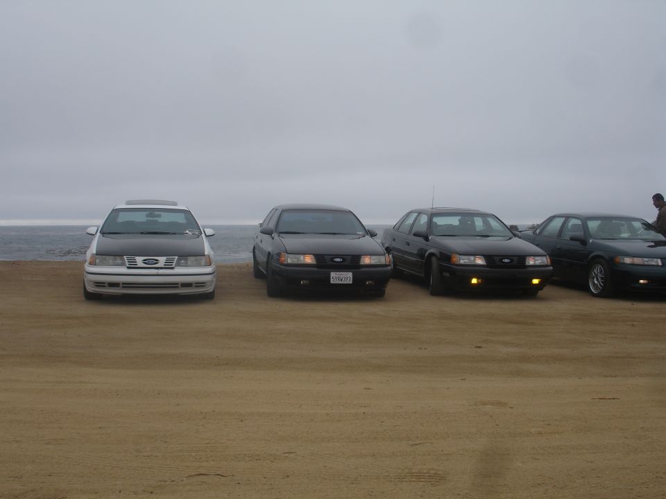 Four sedans parked on a sandy beach with the ocean in the background.