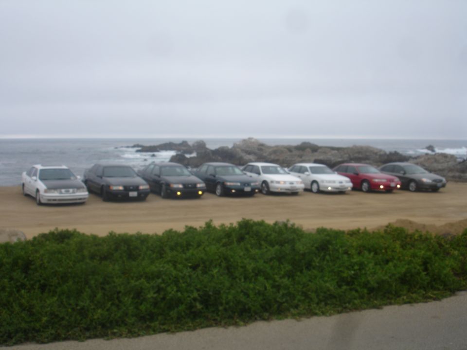 Several sedans parked on a dirt lot by the ocean.