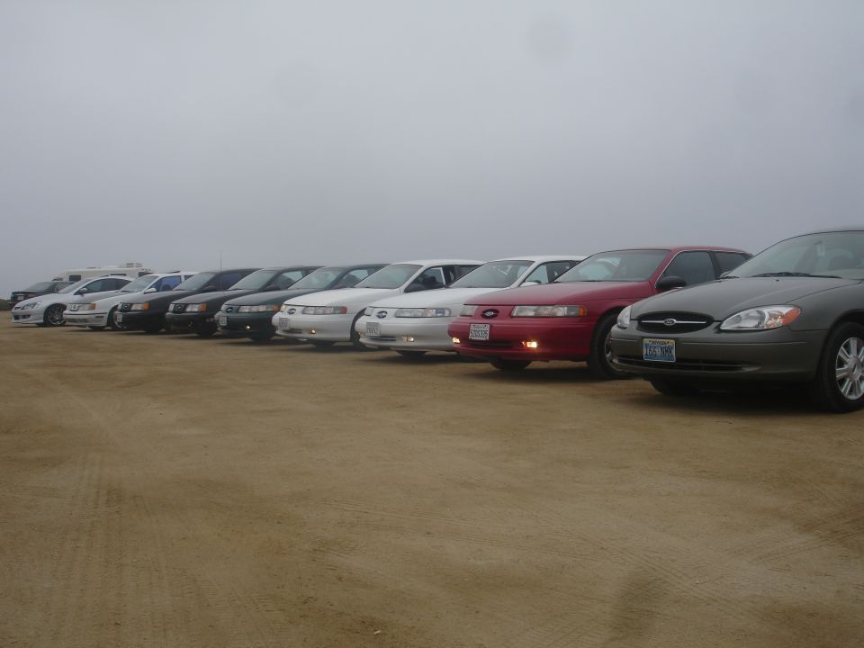 Line of sedans parked on sand, dark gray Ford Taurus in foreground.