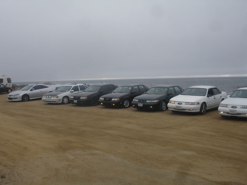 Multiple sedans parked on a beach with the ocean in the background.