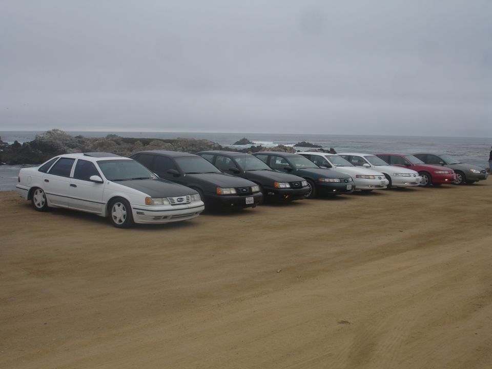 Line of sedans parked on a beach, white sedan with black hood in foreground.