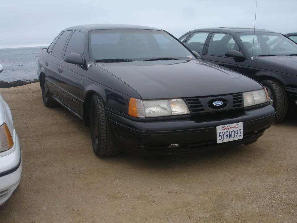 Black 1990 Ford Taurus sedan, front 3/4 view, parked on dirt near ocean.