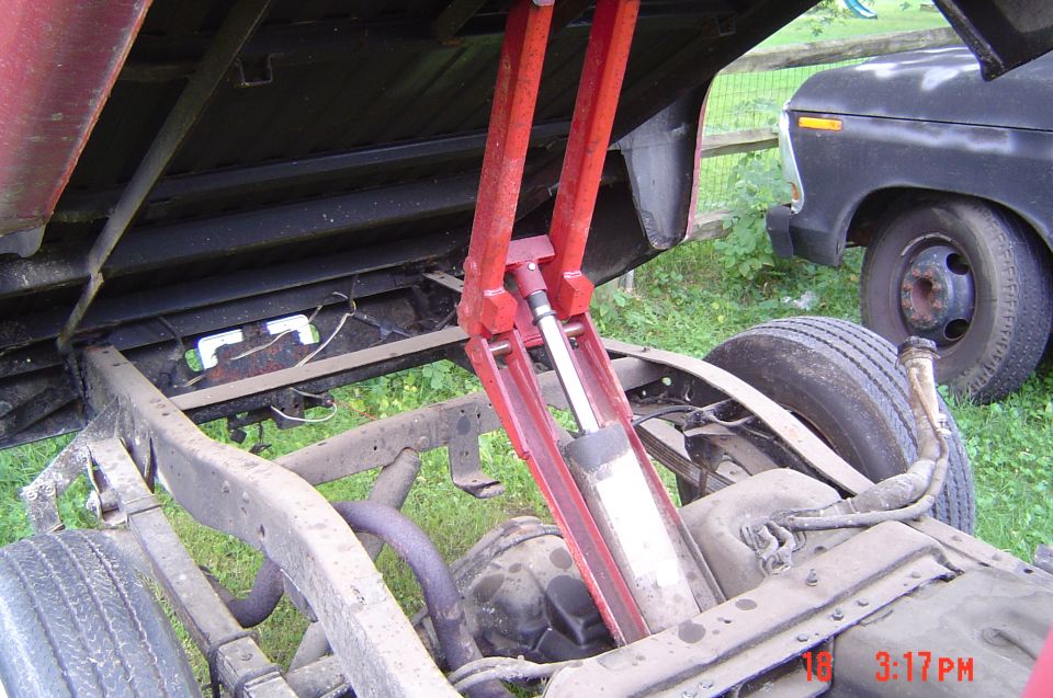 Underside of a red truck bed lifted by a hydraulic hoist, with a black truck in the background.
