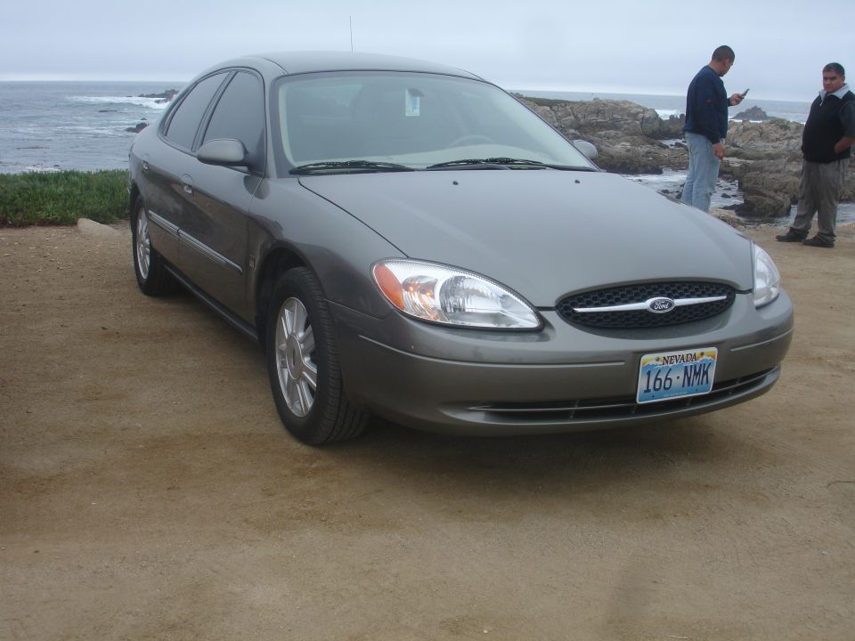 Gray 2000-2007 Ford Taurus sedan parked on dirt lot with ocean and two men in background.