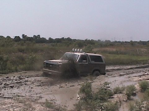 1982 Ford Bronco - Ford Bronco navigating a muddy off-road track, kicking up spray.