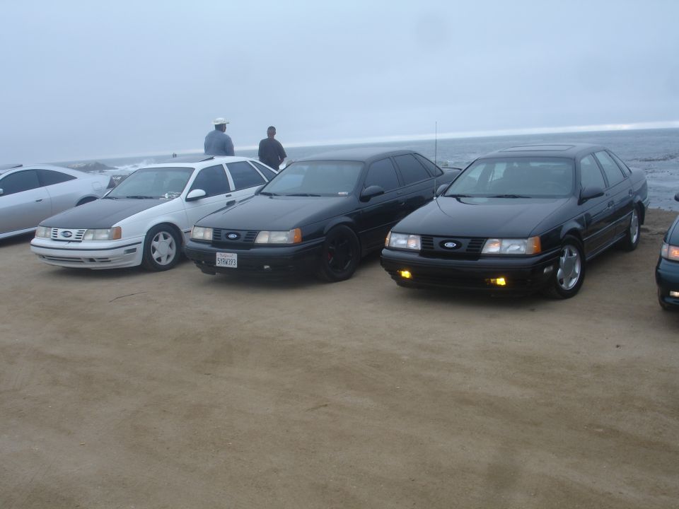 Three Ford Taurus sedans parked on a beach, two black and one white with a black hood.