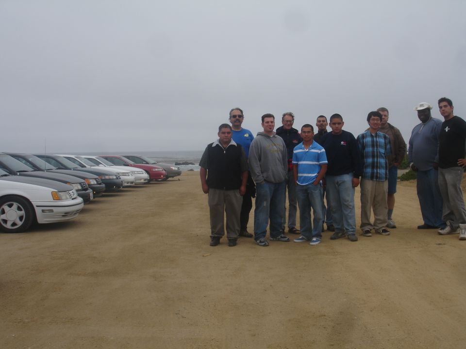 Group of men standing next to a line of sedans parked on a beach.