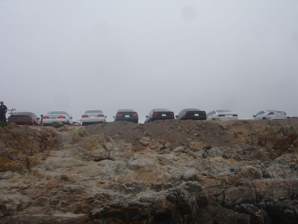 Several sedans parked on a dirt embankment with a rocky shore below and overcast sky.
