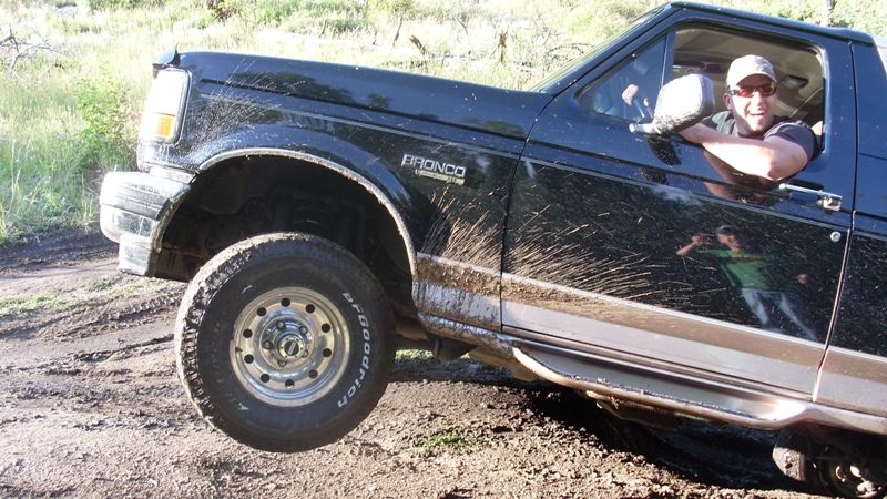 Mud-splattered black Ford Bronco on a dirt trail with driver leaning out window