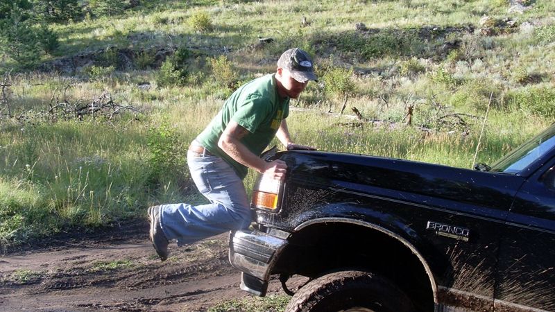 Man leaning on the hood of a black Ford Bronco on a dirt road.